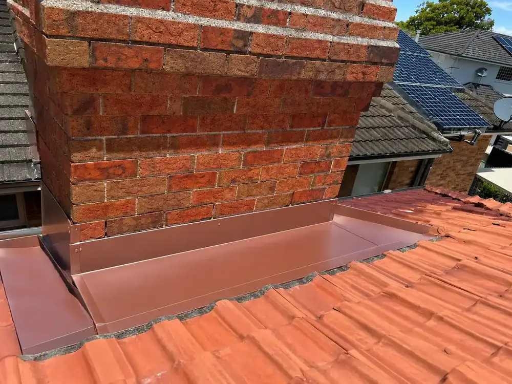 Brown metal chimney apron and side flashings installed around a brick chimney on a terracotta tiled roof in Sydney. 